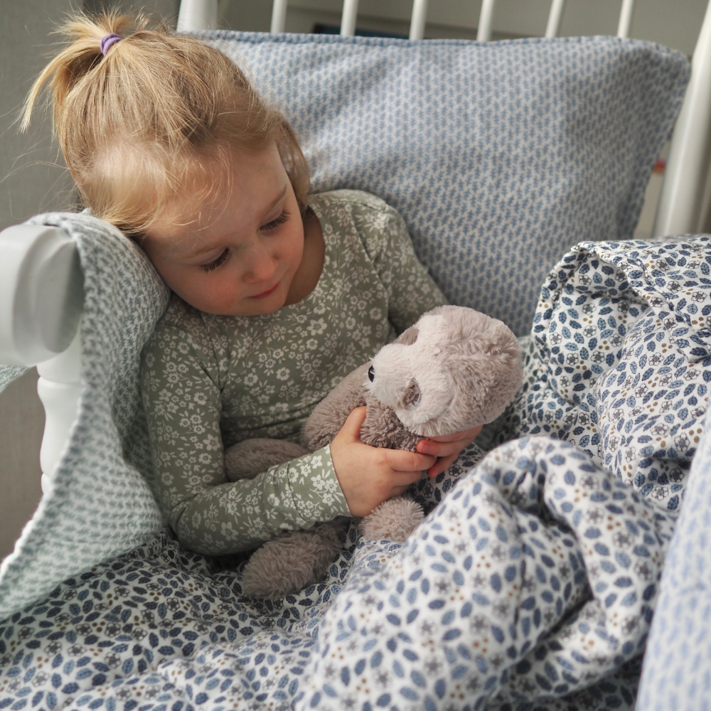 Couverture matelassée pour enfant en mousseline de coton Candide Living de motif Flora en bleu chambray avec une petite fille dans une chaise berçante.