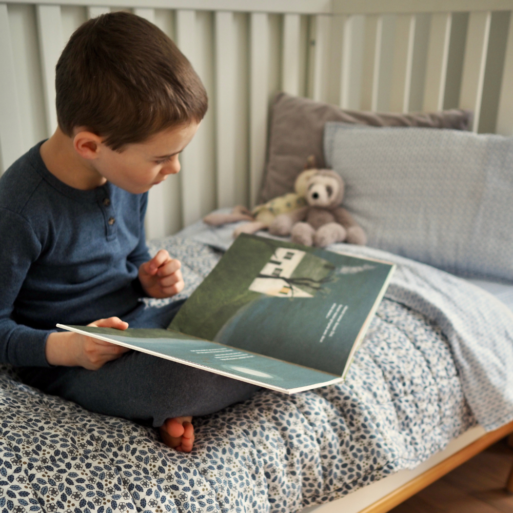 Couverture matelassée pour enfant en mousseline de coton Candide Living de motif Flora en bleu chambray avec un petit garçon regardant un livre sur un lit. 