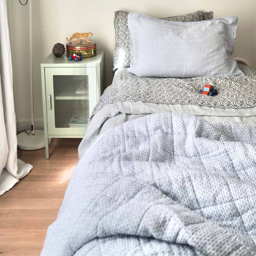 Couverture matelassée pour enfant en mousseline de coton Candide Living de motif Ondine en bleu chambray dans une chambre d'enfant présentée sur un lit simple. 