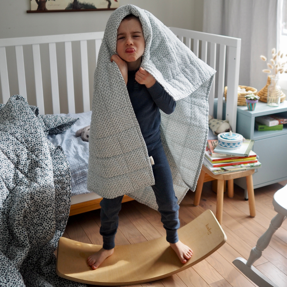 Couverture matelassée pour enfant en mousseline de coton Candide Living de motif Ondine en vert sauge avec un enfant s'amusant dans une chambre. 
