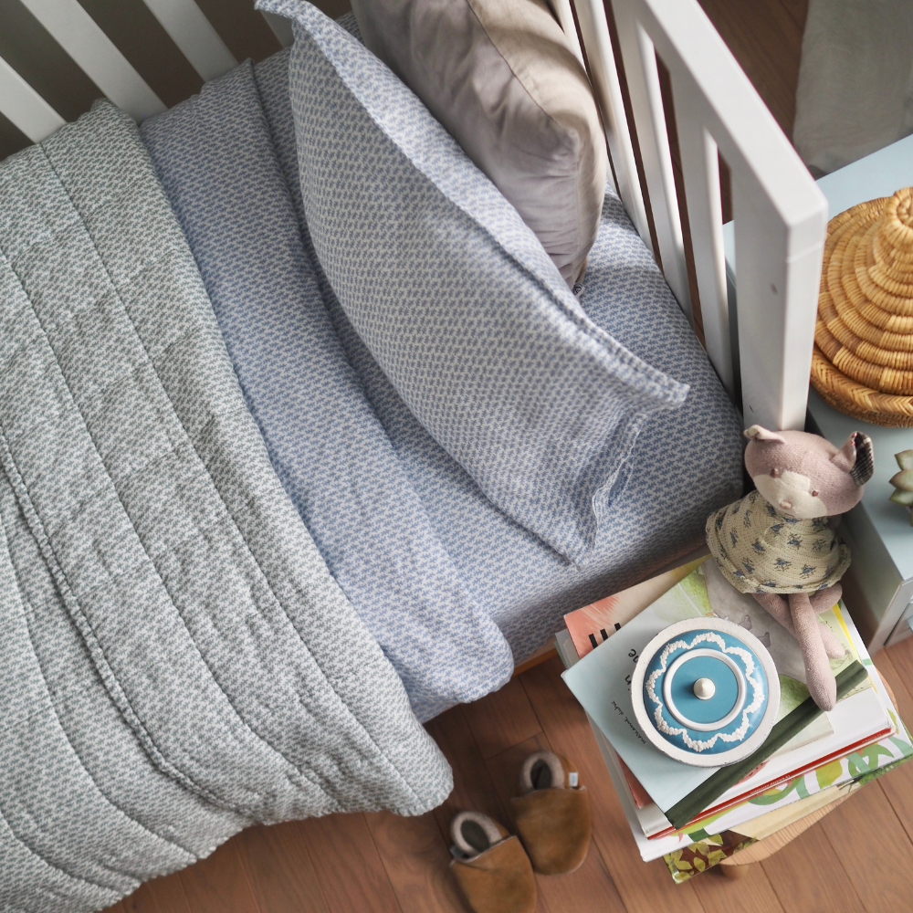 Couverture matelassée pour enfant en mousseline de coton Candide Living de motif Ondine en vert sauge agencée avec l'ensemble de draps Ondine en bleu chambray dans une bassinette. 