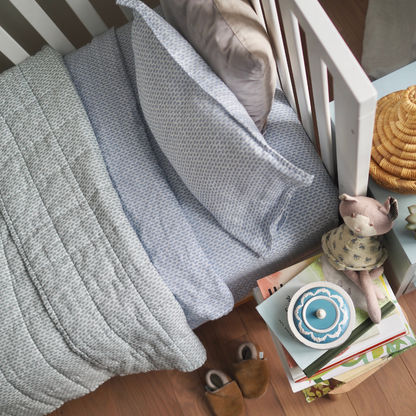 Couverture matelassée pour enfant en mousseline de coton Candide Living de motif Ondine en vert sauge agencée avec l'ensemble de draps Ondine en bleu chambray dans une bassinette. 